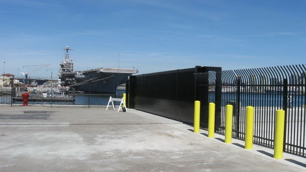 A waterfront security checkpoint featuring a long, black Delta Scientific sliding gate with high visibility yellow bollards, safeguarding a maritime facility with a naval vessel in the background, signifying strong coastal security measures.