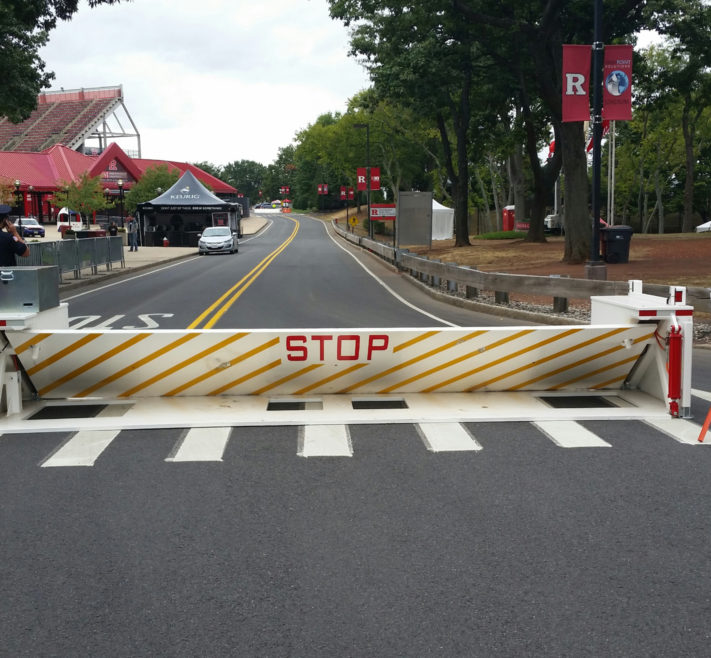 Delta Scientific wedge barrier deployed at university entrance for event security, with directional signage and stadium in the distance, demonstrating perimeter control solutions for campus safety.