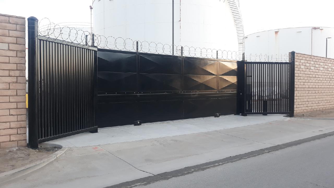 Robust black Delta Scientific sliding security gate installed at a facility entrance, complemented by reinforced walls and topped with concertina wire, symbolizing high-level perimeter defense against unauthorized access.