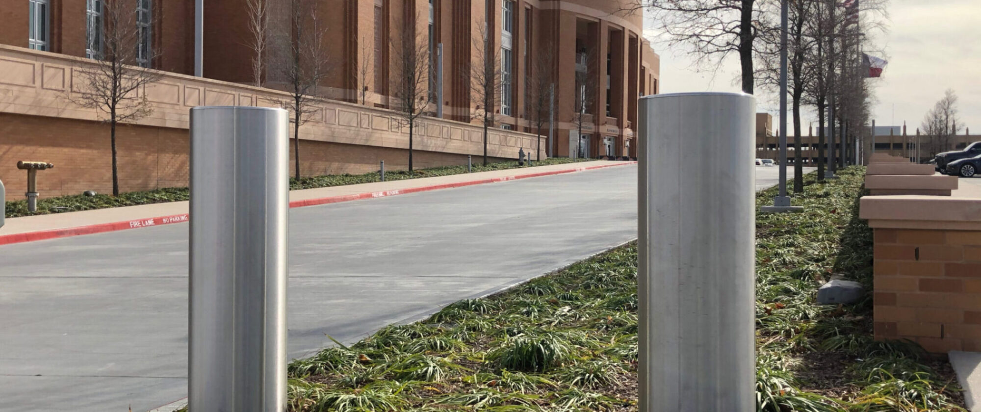 Delta Scientific bollards installed at a facility entrance for secure access control, with an architectural brick building in the background, a clear sky, and the American flag waving in the distance, symbolizing robust security in a professional setting.