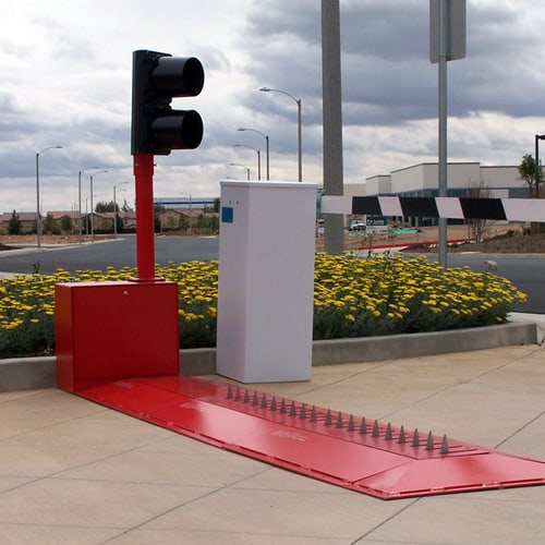 Red Delta Scientific surface-mounted spike barricade in the deployed position with a traffic control signal post, designed for high-security vehicle access points to enforce one-way traffic flow and prevent unauthorized entry.