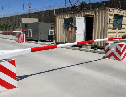 Red and white Beam Barricade TT212E arm blocks a paved road near a tan portable building with a door open, barbed wire fencing, and equipment in the background.