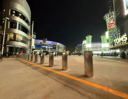 Nighttime view of an urban plaza with illuminated buildings and Bollard DSC720 units lining the walkway. Scattered people move in the distance, while signs for “TOM'S WATCH BAR” and “Crypto.com Arena” add a lively atmosphere.