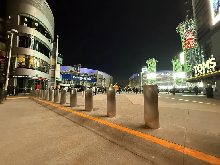 Nighttime view of an urban plaza with illuminated buildings and Bollard DSC720 units lining the walkway. Scattered people move in the distance, while signs for “TOM'S WATCH BAR” and “Crypto.com Arena” add a lively atmosphere.