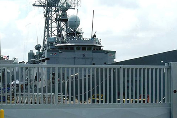 Heavy-duty Delta Scientific security gates guard the entrance to a naval port, with a military ship visible in the background, illustrating Delta's strategic importance in securing critical maritime infrastructure.