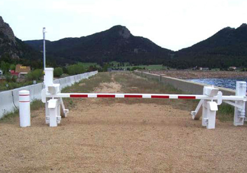 Delta Scientific barrier arm system securing a water reservoir, with a prominent red and white arm creating a checkpoint on a gravel road in a scenic mountainous area, reflecting the company's role in safeguarding critical public water supplies.