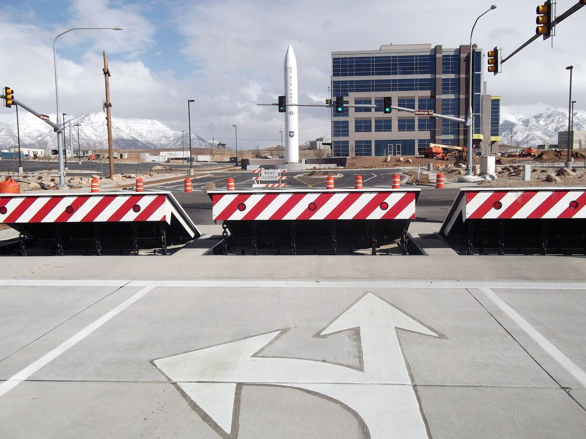 Roadway entrance with three Delta Scientific barricades displaying diagonal red and white stripes and 'ROAD CLOSED' signage, set against a backdrop of snow-capped mountains and a modern office building, illustrating the deployment of certified traffic control solutions to enhance safety.
