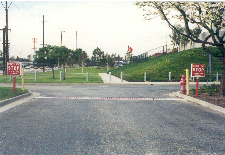 Security checkpoint with a Delta Scientific vehicle barrier arm raised, tire spikes embedded in the road surface, and warning signs indicating the wrong way, signifying a well-guarded entrance with an American flag fluttering in the background.