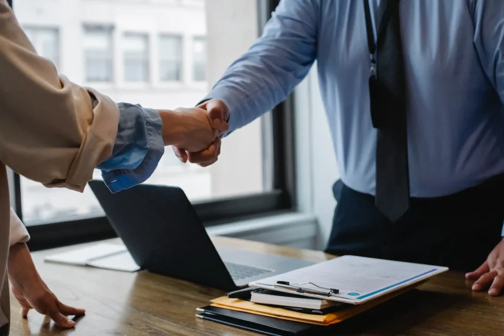 Two professionals engage in a firm handshake above a wooden table with work documents and a laptop, symbolizing a partnership or agreement, reflecting Delta Scientific's commitment to professional collaborations and security solutions contracts.