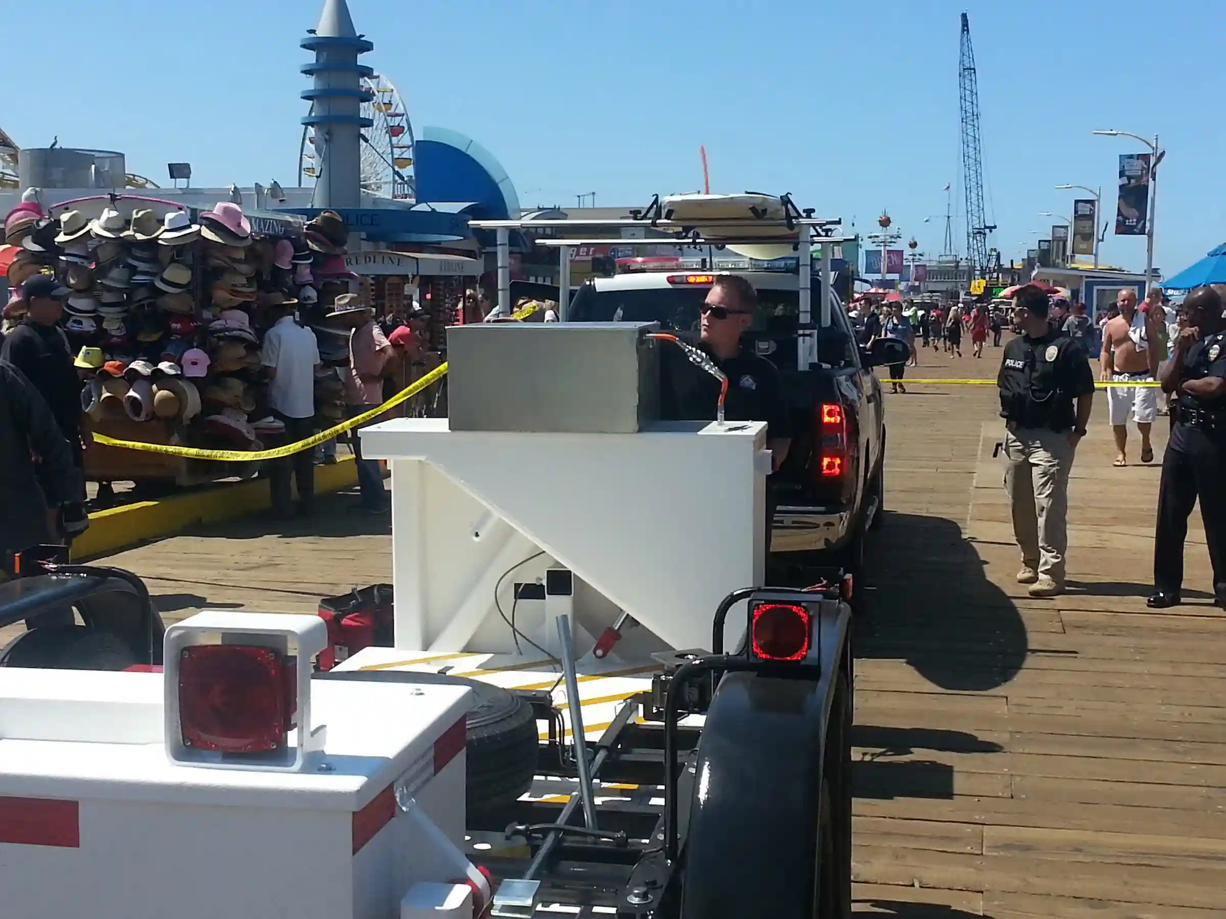 A Delta Scientific mobile barrier deployed on a lively boardwalk, with police oversight ensuring the safety of the bustling public space filled with visitors and vendors near ocean-side attractions.