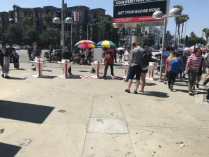 People gather around registration tables outside a convention center on a sunny day. Event Security monitors the area, while banners advertising badge pickup and portable vehicle barriers help guide attendees safely. Some hold umbrellas for shade.