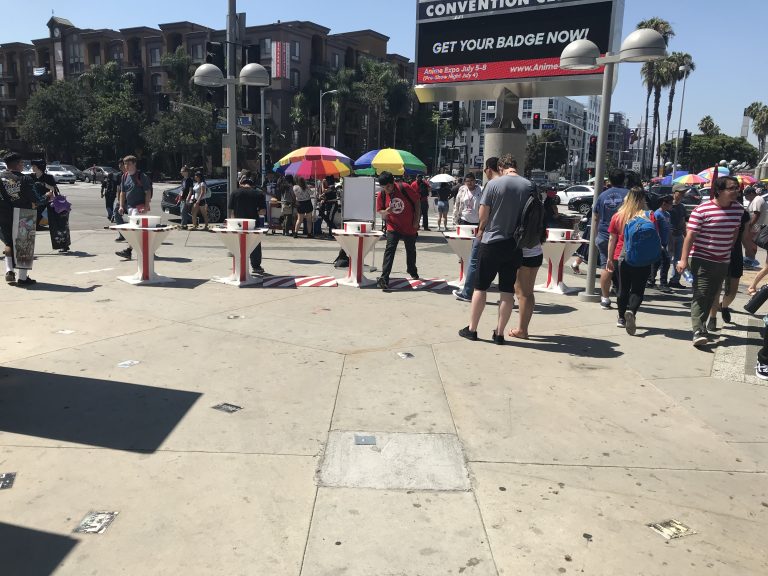People gather around outdoor check-in tables under umbrellas at a busy street corner near a convention center, where event security and portable vehicle barriers ensure safety on a sunny day.
