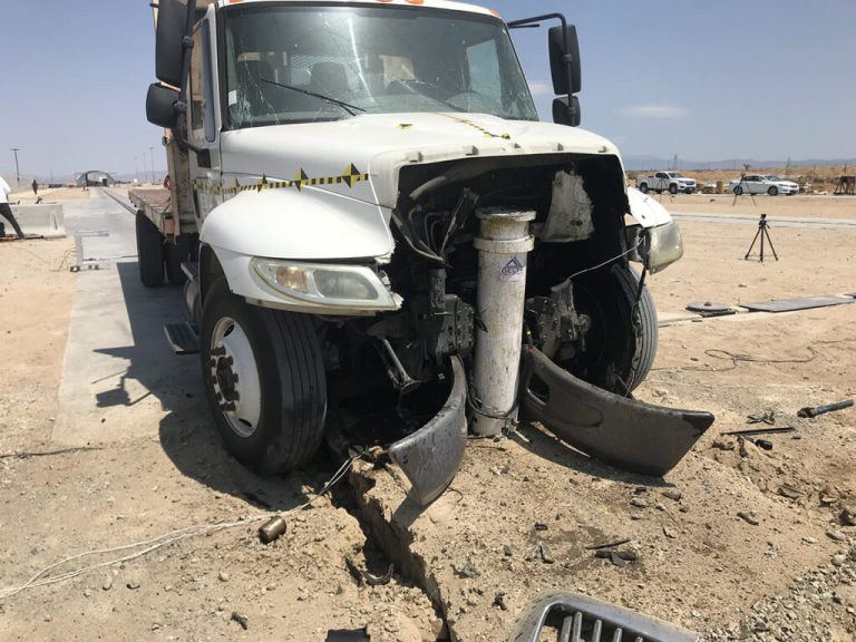 A white truck with severe front-end damage is crashed into a large metal post and bollards in a desert area, with debris scattered on the ground.