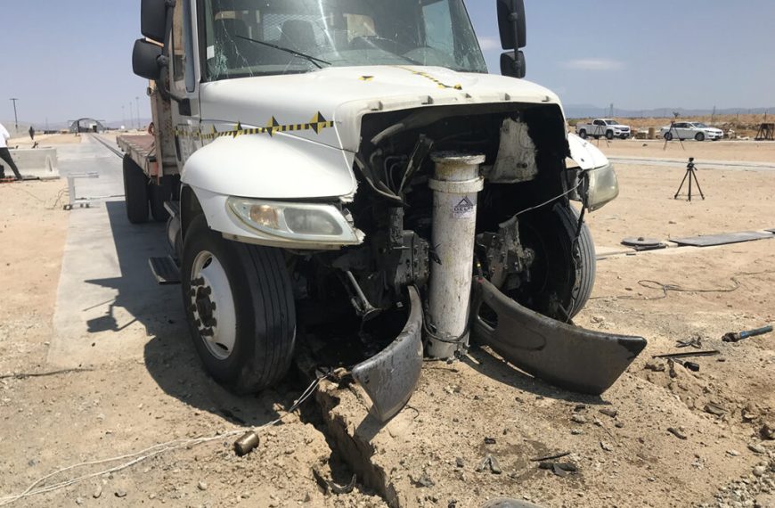 A white truck with severe front-end damage is crashed into a large metal post and bollards in a desert area, with debris scattered on the ground.