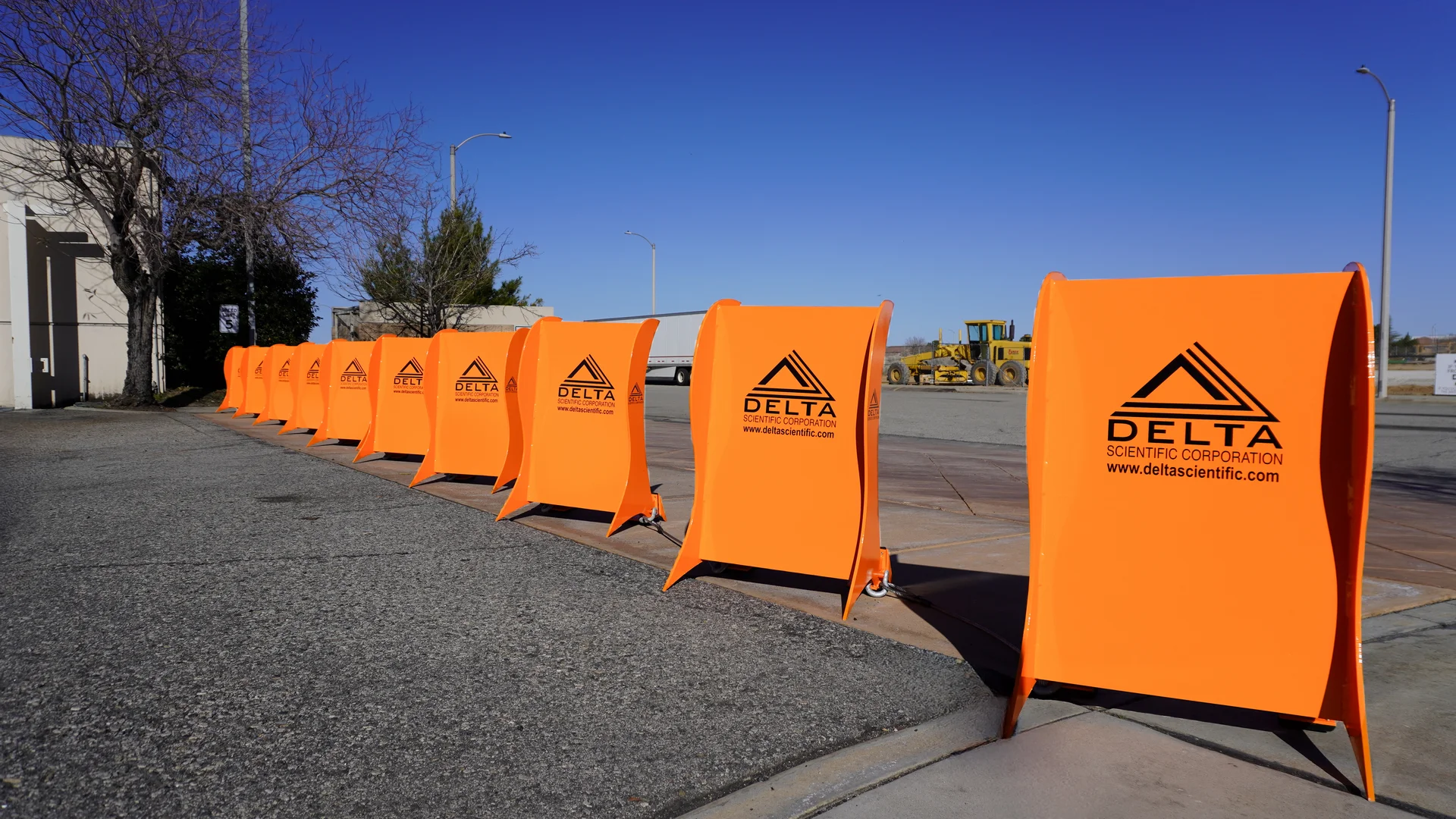 A row of bright orange Delta Scientific barricades is lined up on a sidewalk next to a street, providing crowd protection, with construction equipment visible in the background.