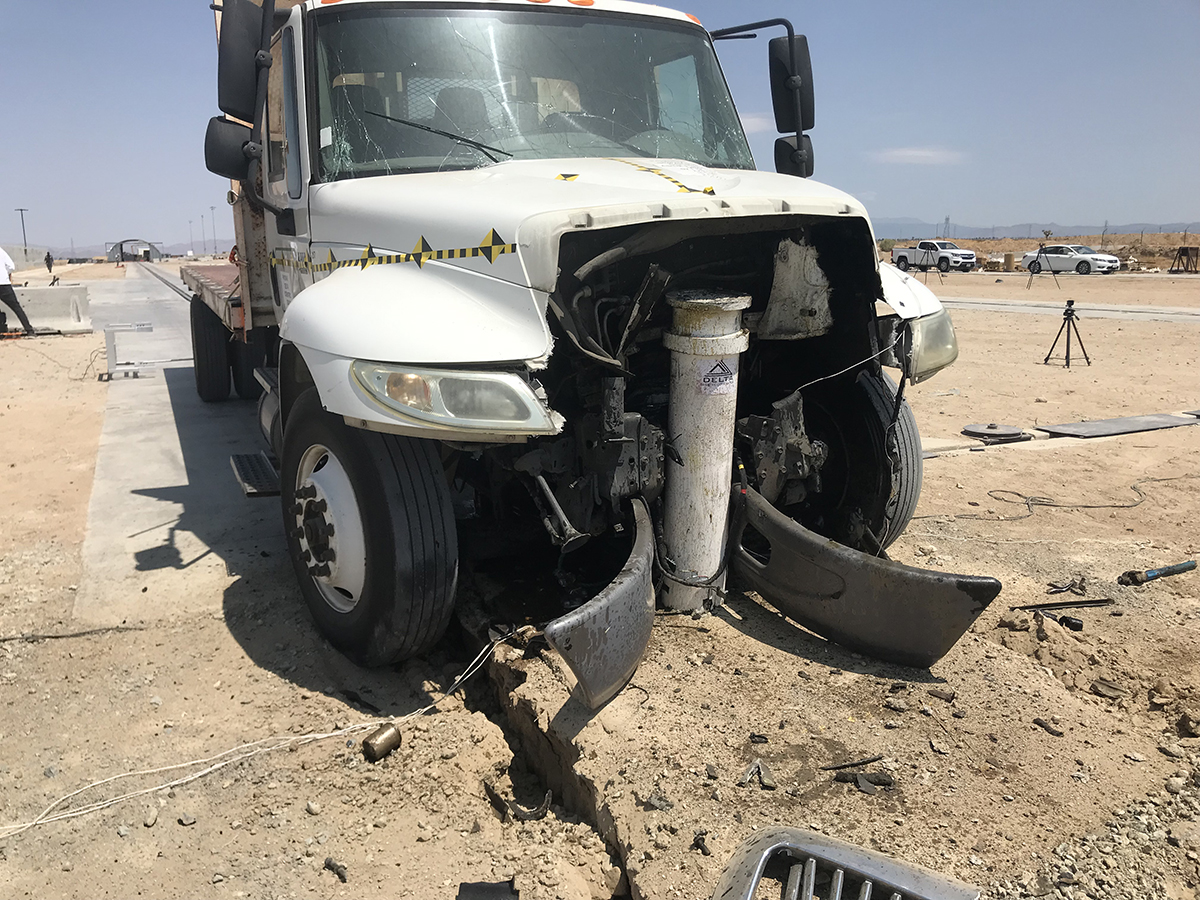 A white truck with severe front-end damage is stopped on a cracked concrete surface; crash-rated bollards could have offered better protection than the barrier now lodged under its hood—something facility owners should consider.