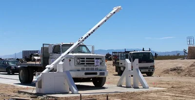 A white truck is stopped at a raised security barrier gate in a desert area, with other vehicles and construction equipment in the background.