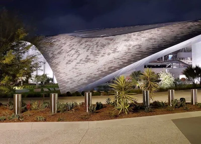 A modern building with a curved, textured facade at night, surrounded by landscaped plants and illuminated by outdoor lighting.