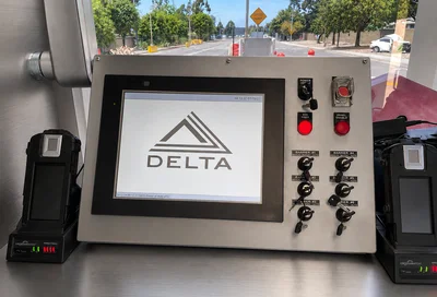 A control panel with a touchscreen displaying the DELTA logo, several switches, buttons, and handheld devices, set inside a vehicle with a road visible through the window.