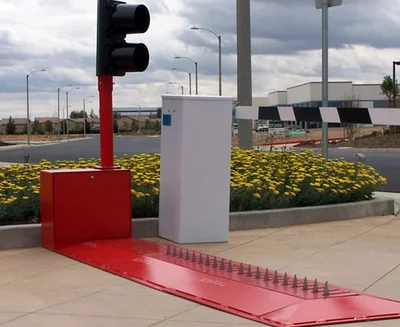 A red traffic control barrier with metal spikes blocks a driveway entrance near a traffic light and a white control box, with yellow flowers in the background.