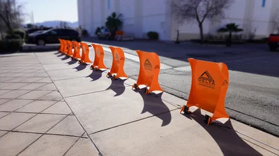 A row of bright orange plastic barriers lines the edge of a sidewalk next to a street on a sunny day.