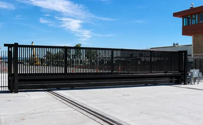 A large black metal sliding security gate is installed at the entrance of an industrial or commercial facility on a sunny day.
