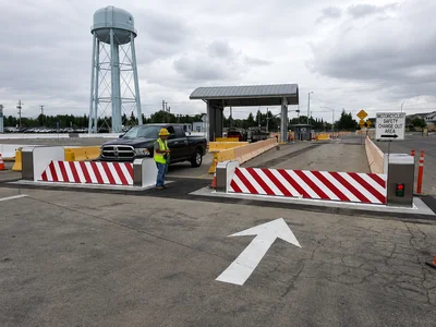 A security checkpoint with a raised barrier, a pickup truck, a worker in a safety vest and helmet, and a water tower in the background.