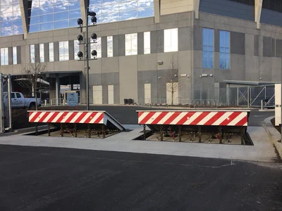 Two red-and-white striped vehicle barriers are installed in the ground outside a modern building, with a blacktop road in the foreground.