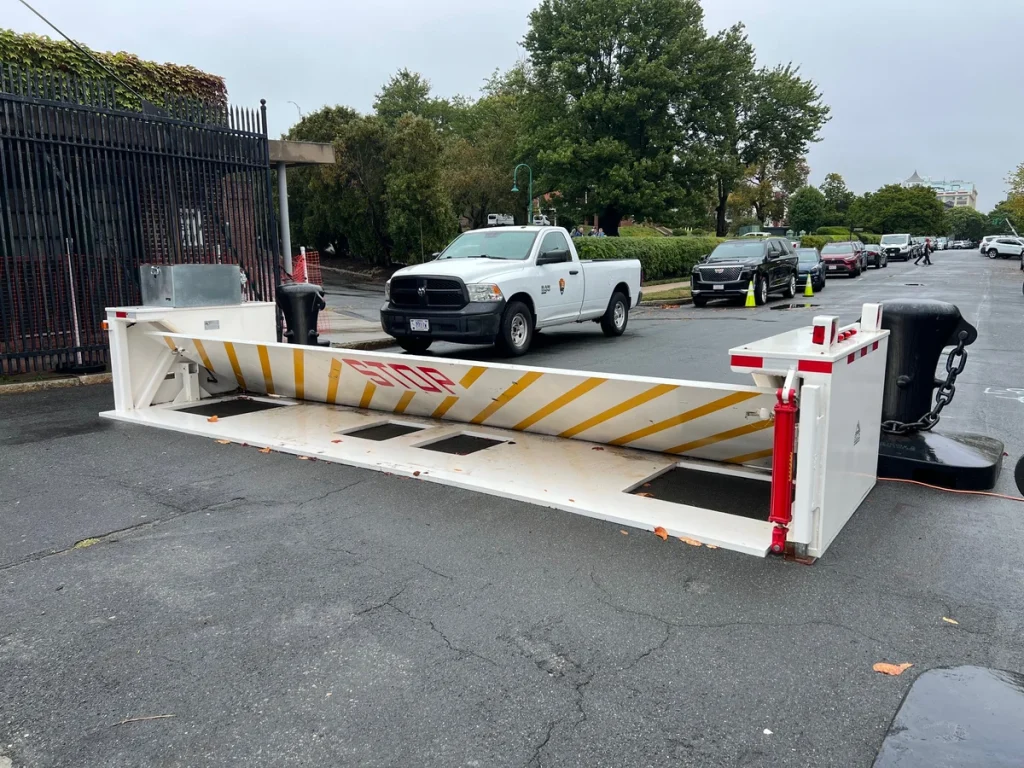 A white security barrier with red "STOP" text is raised on a street, blocking vehicle access. This type of vehicle barrier enhances security by preventing unauthorized entry. A white pickup truck and several cars are in the background.