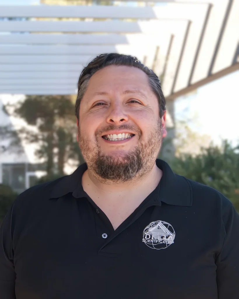 A man with a beard, wearing a black polo shirt with a Beam Barricades logo, smiles outdoors under a white pergola structure.