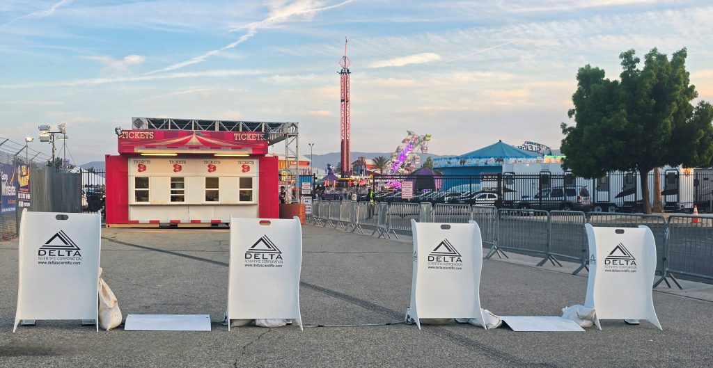 A row of crowd protection barriers blocks an entrance to a fairground with a red ticket booth and carnival rides in the background, ensuring effective crowd control often necessary in busy cities.