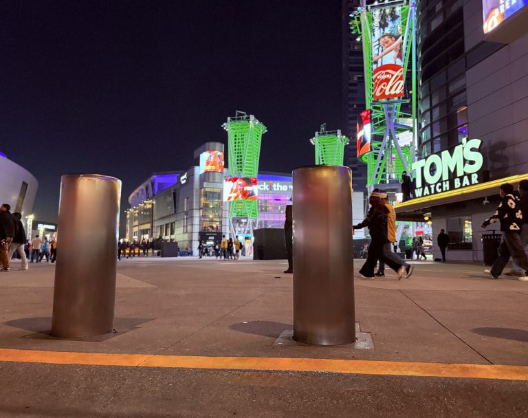 Street view at night in bustling cities showing metal bollards and crowd protection barriers in the foreground, people walking, and illuminated buildings with ads and signs including TOM’S WATCH BAR and Coca-Cola glowing in the background.