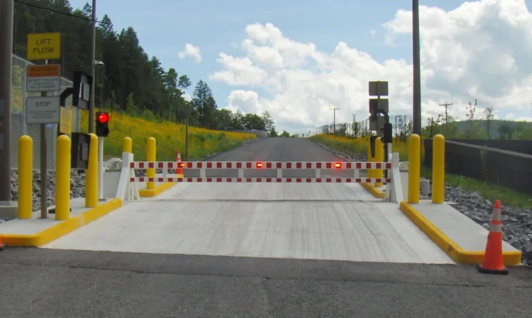 A closed railroad crossing gate with red lights, yellow posts, and warning signs blocks a paved road on a sunny day, serving as one of the essential security barriers designed to ensure safety at train crossings.