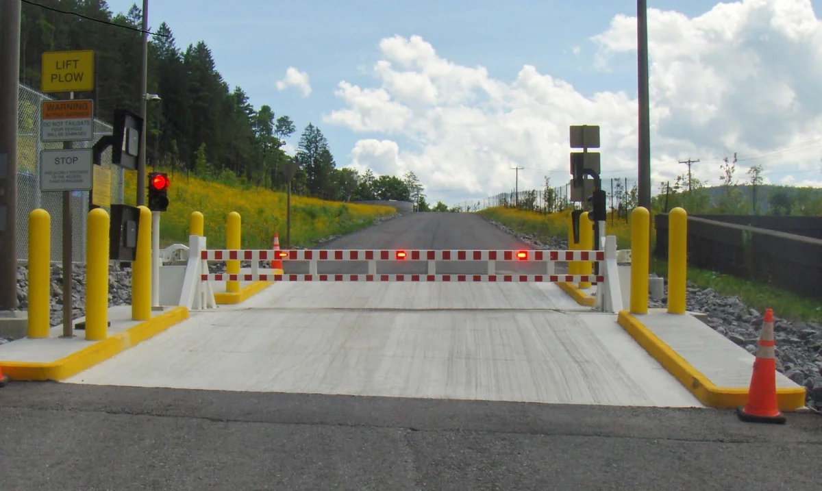 A closed railroad crossing gate with red lights, yellow posts, and warning signs blocks a paved road on a sunny day, serving as one of the essential security barriers designed to ensure safety at train crossings.