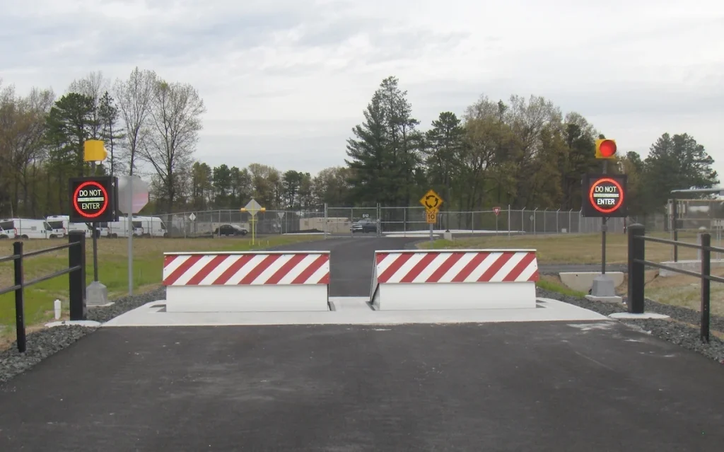 A secured road entrance is blocked by two raised, crash-rated barriers featuring “Do Not Enter” signs and red lights on both sides; trees and fencing are visible in the background.