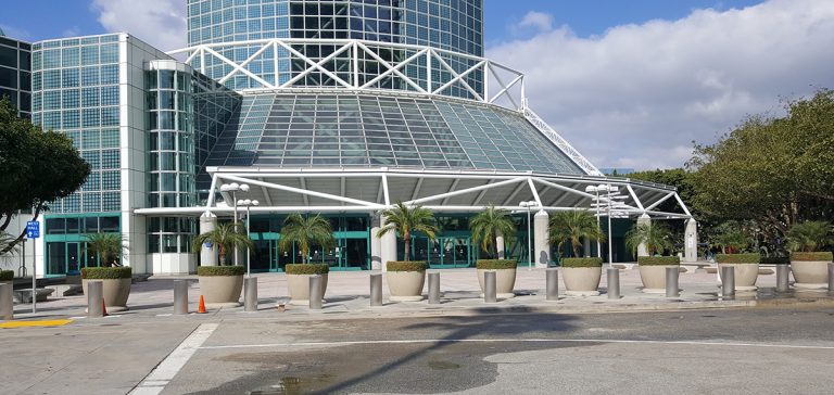 Glass-fronted convention center entrance with white structural beams, large windows, and potted palm trees lined up outside on a sunny day—protected discreetly by M30 bollards designed to enhance safety while maintaining the venue’s welcoming aesthetic.