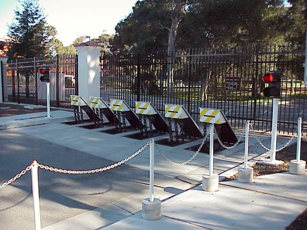 Five hydraulic vehicle barriers with yellow hazard signs are raised in front of a gated entrance, next to a security fence and warning lights, illustrating the practical application of Shallow vs. Deep Foundation Vehicle Barriers in high-security environments.
