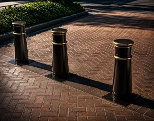 Three black bollards with gold-colored trim stand in a row on a red brick sidewalk as architectural design elements.