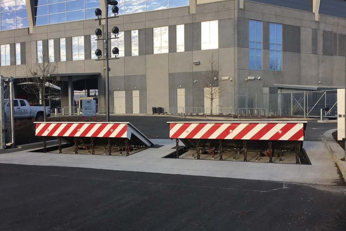 Two raised security barrier gates with red and white diagonal warning stripes installed in the ground in front of a stadium building.
